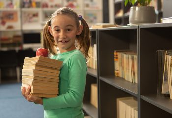 Side view of smiling Caucasian schoolgirl with books and apple standing in library at elementary school